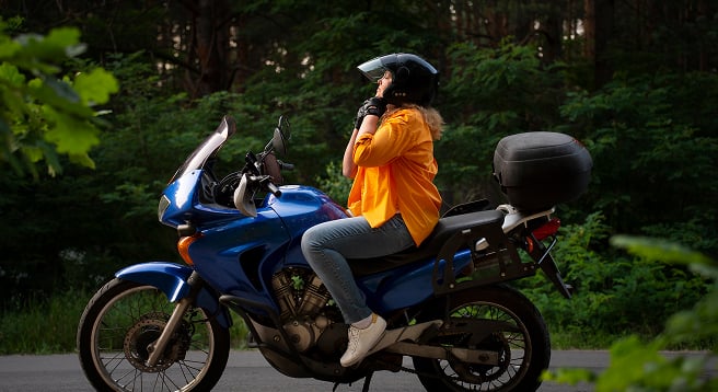 Orman yolunda mavi motosikleti üzerinde sürüş güvenliği için siyah kaskını takan kadın sürücü.