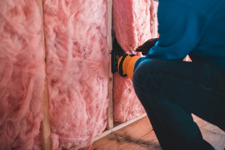 Pembe cam yünü yalıtımlı ahşap duvarda elektrik tesisatı çalışması yapan eldivenli işçi.