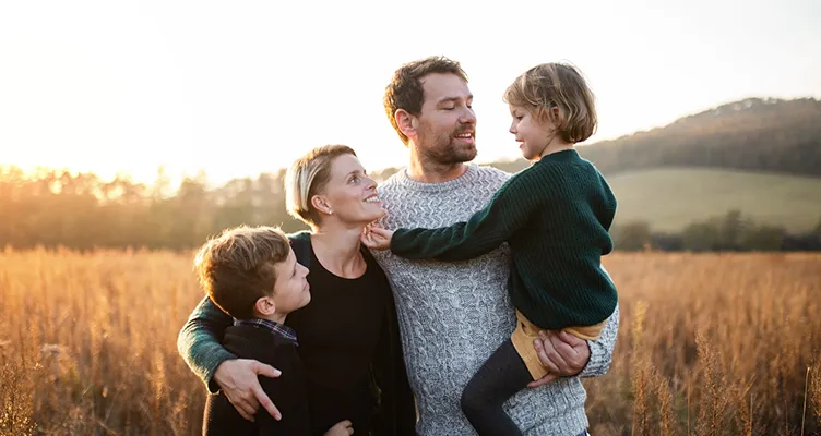 Allianz güvencesiyle gün batımında doğada huzurla vakit geçiren, birbirine sevgiyle bakan anne, baba ve çocuklu aile.