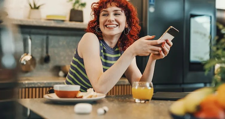 Mutfakta akıllı telefonuyla Allianz dijital hizmetlerini kullanan gülümseyen kızıl saçlı kadın.