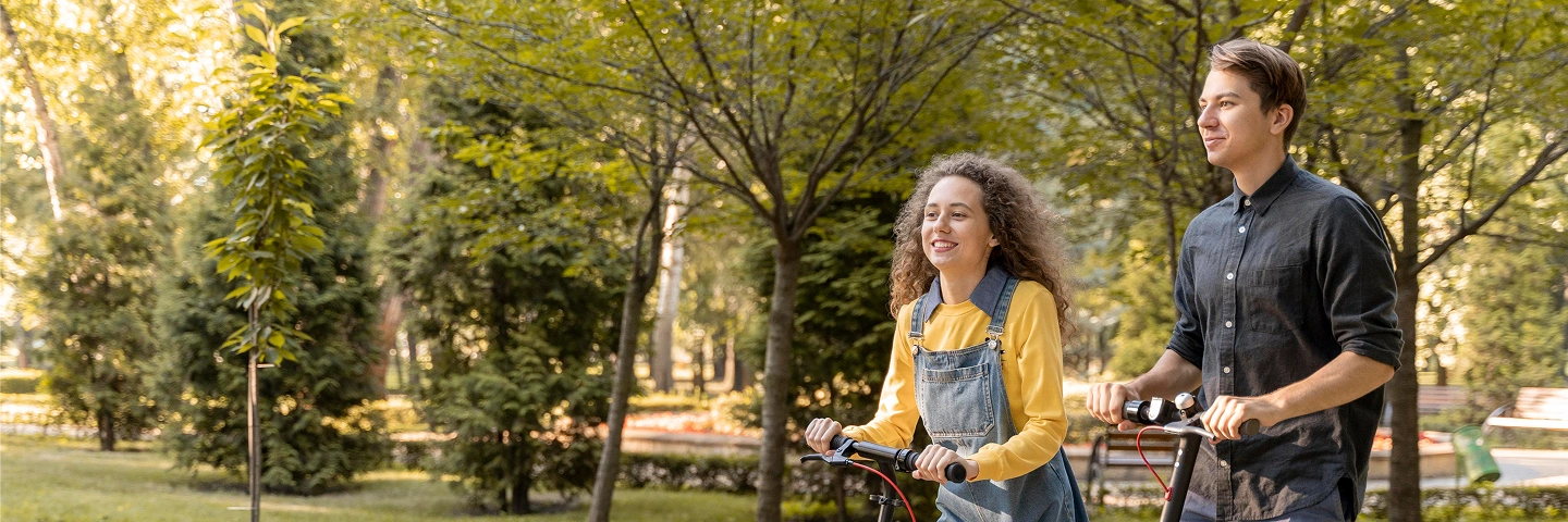Güneşli parkta elektrikli scooter kullanan, güvende ve mutlu hisseden genç çift.
