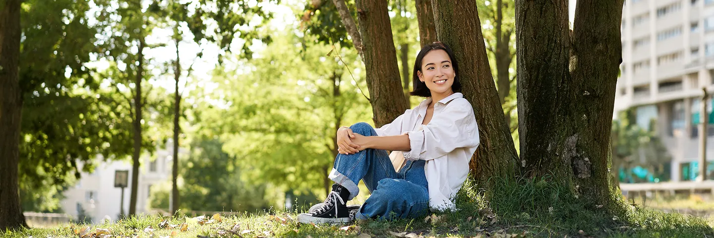 Parkta büyük bir ağaca yaslanarak çimlerde oturan ve huzurla gülümseyen genç kadın.