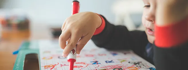 Küçük bir çocuk elinde kırmızı kalemle alfabe çalışma kitabında harfleri yazarak öğreniyor.