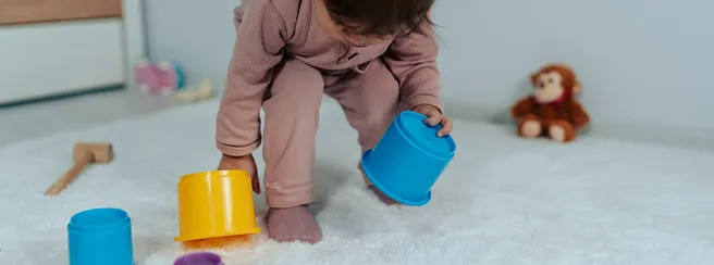 Pembe kıyafetli bir bebek, beyaz tüylü bir halı üzerinde renkli oyuncak bardaklarla oynuyor.
