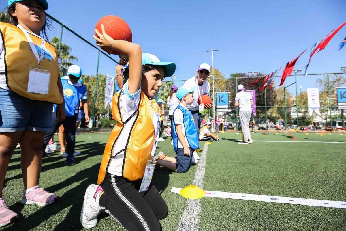 Sarı yelekli bir kız çocuğu, açık havada diz çökmüş, turuncu bir topu fırlatmaya hazırlanırken, diğer çocuklar ve antrenörler spor etkinliğinde yer alıyor.