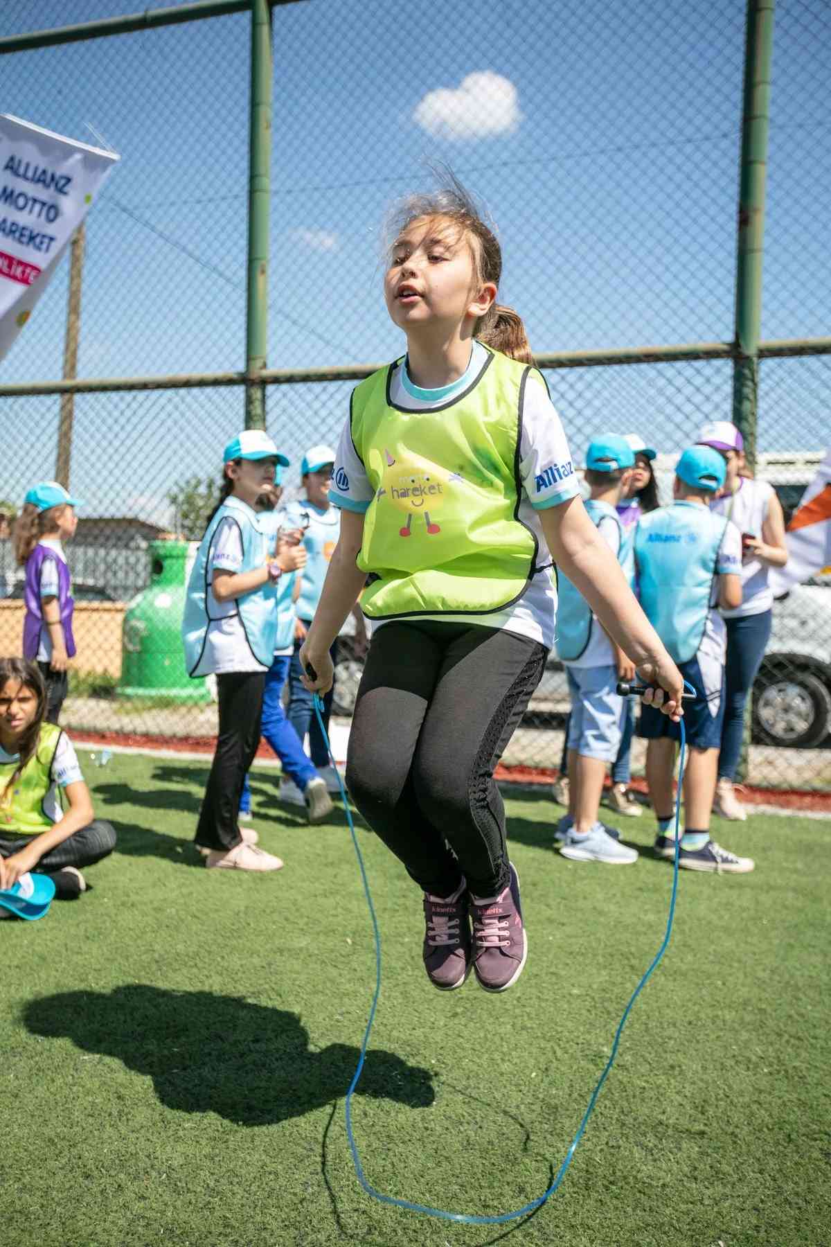 Sarı yelekli, enerjik bir kız çocuğu güneşli çim sahada ip atlarken görülüyor. Allianz etkinliğinde spor ve hareket.