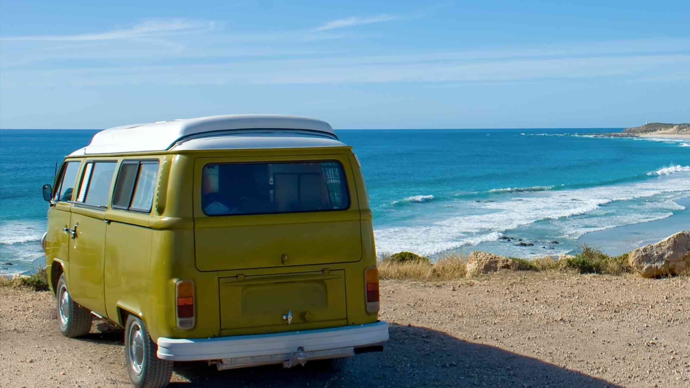 A yellow caravan parked on a road overlooking the sea. A moment of free travel with the blue sky, turquoise ocean waves, and coastline.