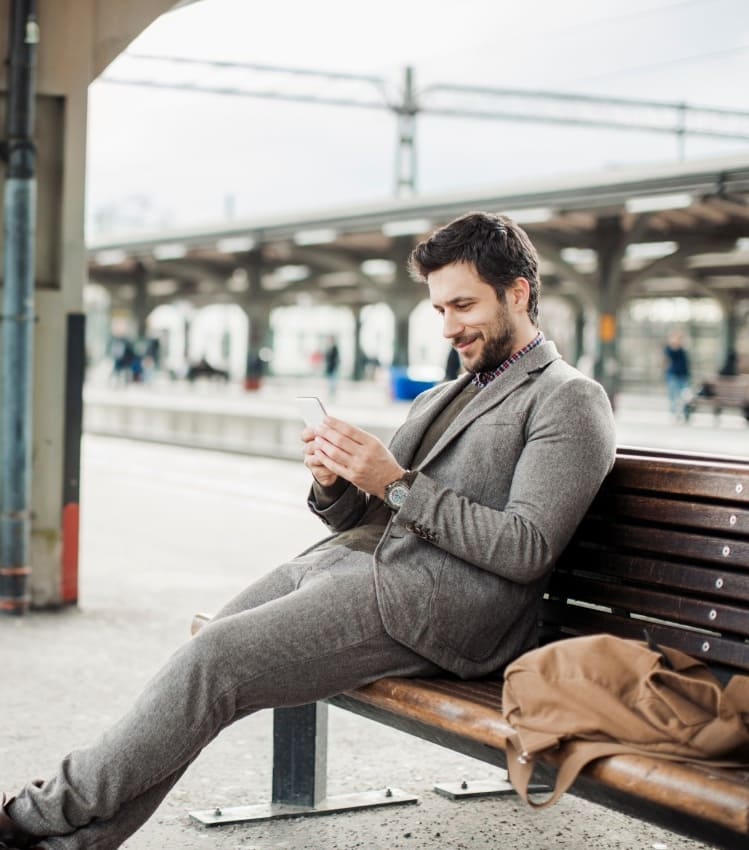 Tren istasyonunda bankta oturmuş, akıllı telefonuna bakarak gülümseyen şık bir adam ve yanındaki çantası.