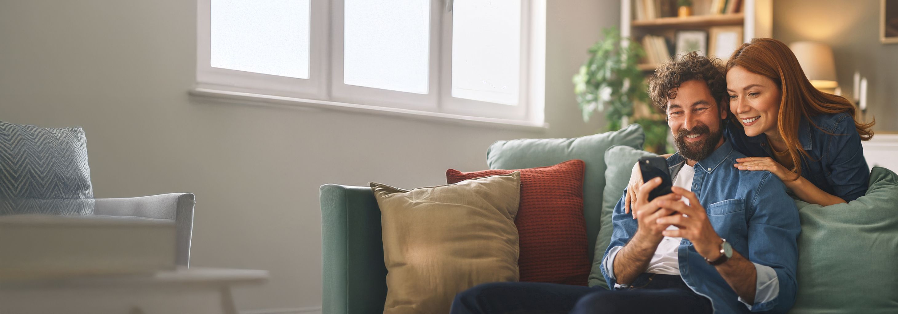 Evde kanepede otururken akıllı telefondan Allianz mobil işlemlerini inceleyen ve gülümseyen mutlu çift.