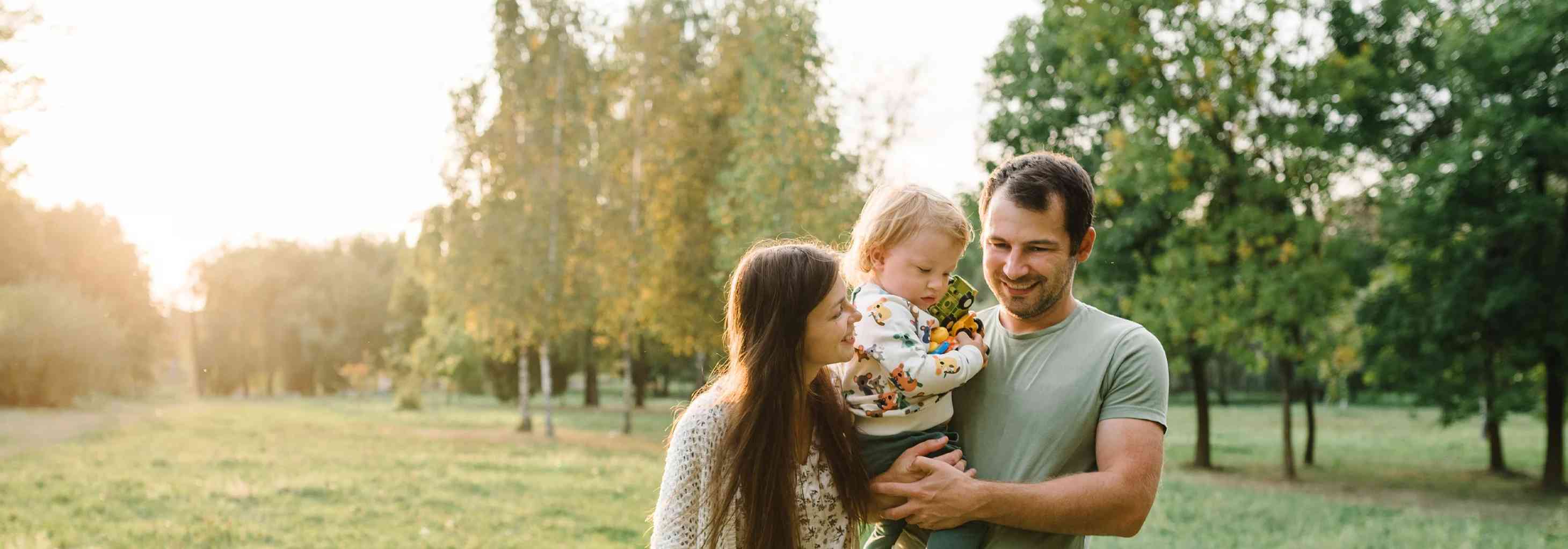 Güneşli bir parkta kucağında çocuğuyla gülümseyen anne ve baba, aile mutluluğu ve huzuru.