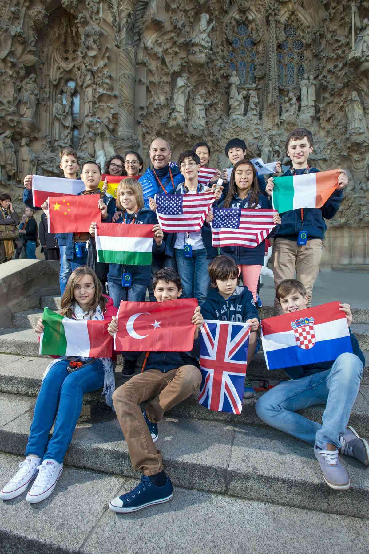 Çeşitli ülkelerden gençler ve bir yetişkin, tarihi bir yapının önünde ulusal bayraklarını tutarak gülümsüyor.