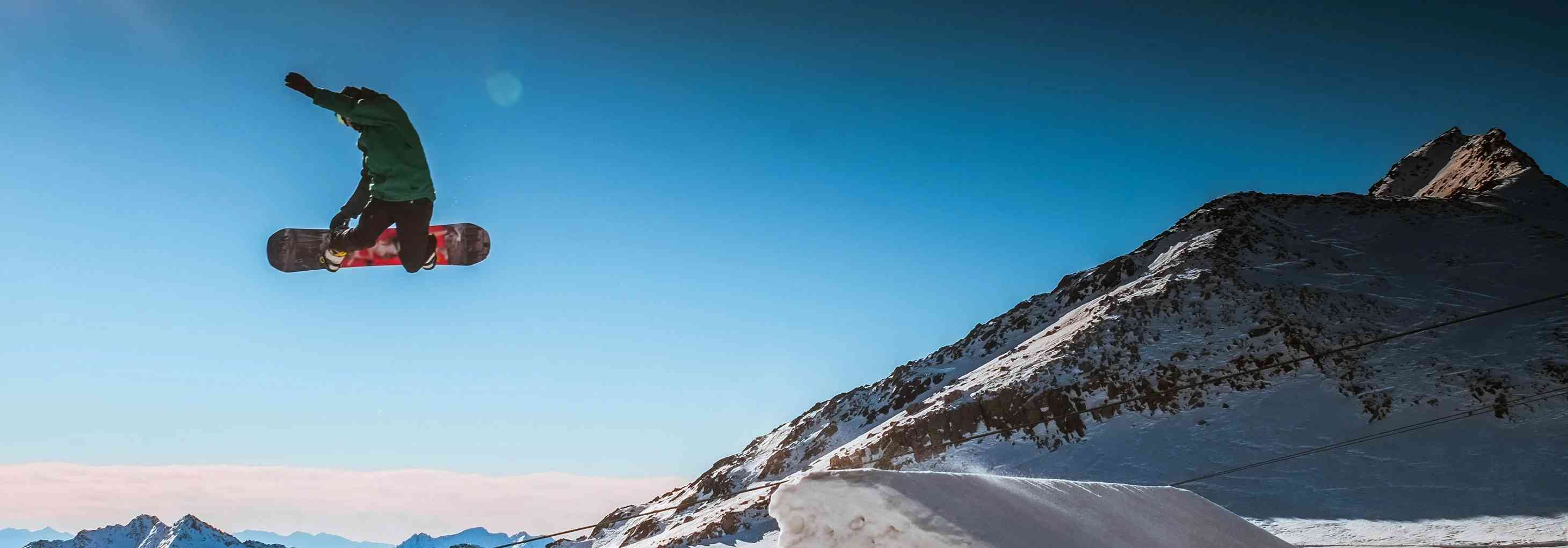 Bir snowboardcu karda zıplarken havada. Karlı dağlar ve mavi gökyüzü arka planda. Allianz spor sponsorluğu.