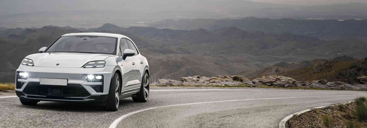 Virajlı dağ yolunda ilerleyen beyaz Porsche SUV, gri dağ manzarası eşliğinde güvenli ve modern bir sürüş deneyimi sunuyor.