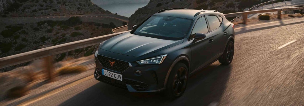 Mat siyah bir Cupra SUV'nin virajlı dağ yolunda dinamik sürüşü, arka planda deniz ve yemyeşil tepeler. Güvenli yolculuk.