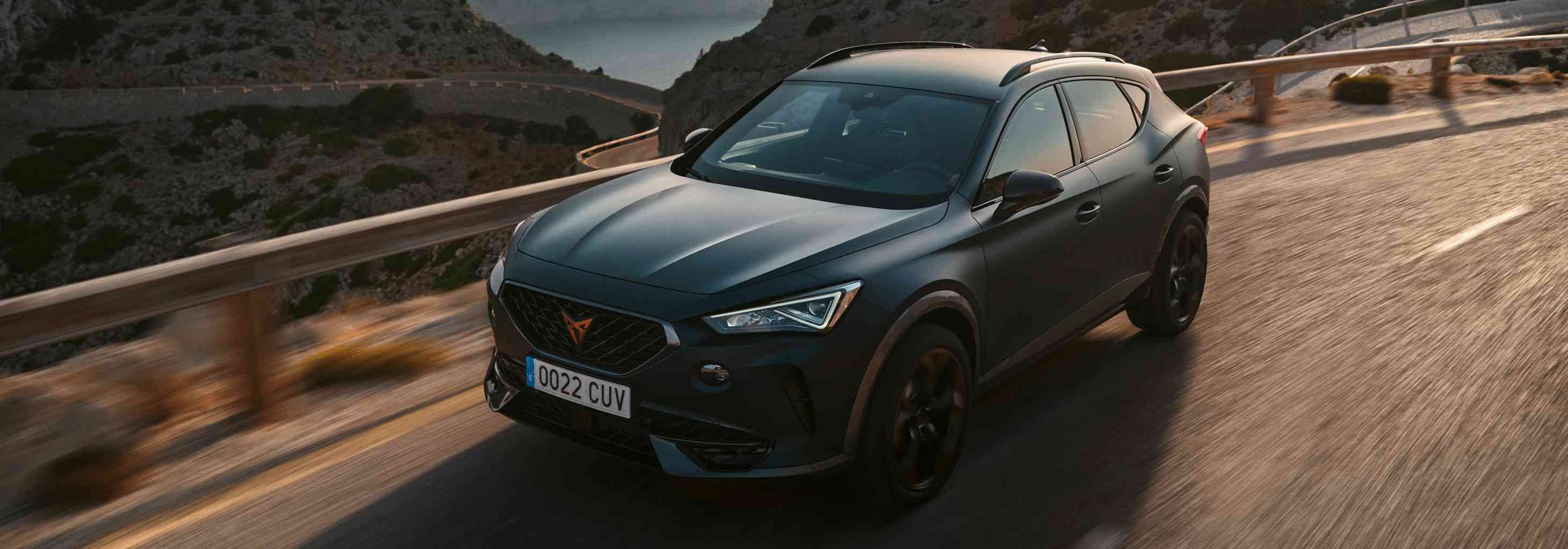 Mat siyah bir Cupra SUV'nin virajlı dağ yolunda dinamik sürüşü, arka planda deniz ve yemyeşil tepeler. Güvenli yolculuk.