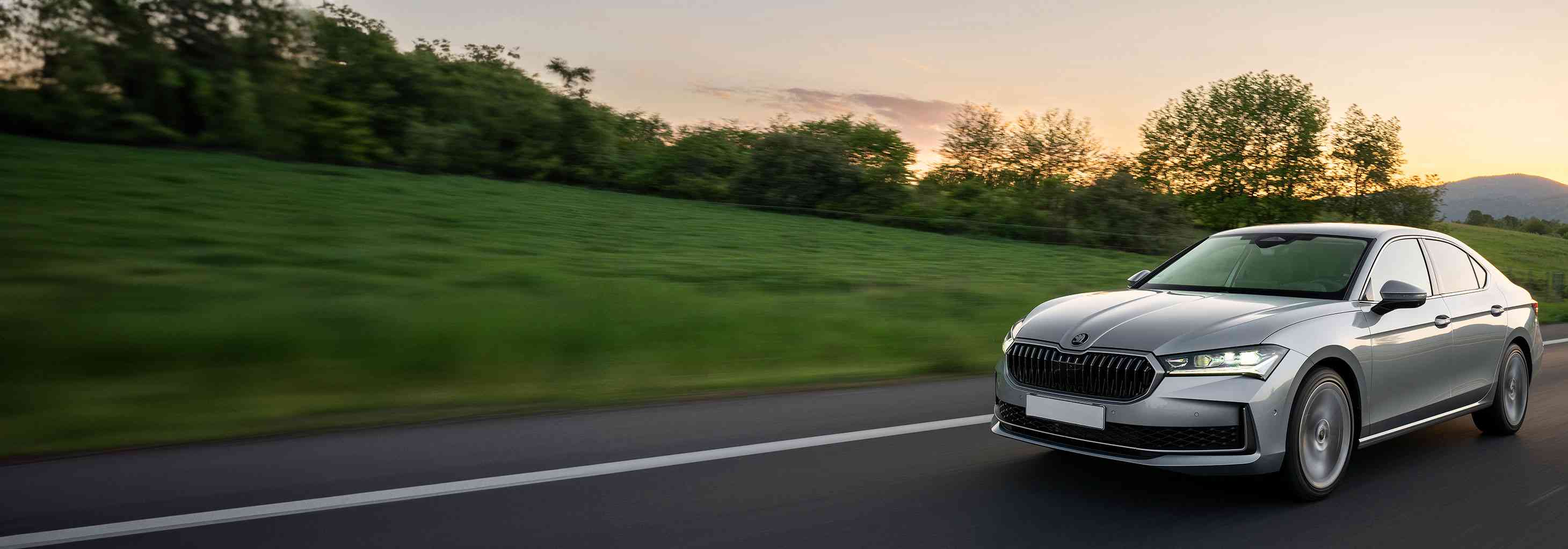 Gün batımı ışıkları altında yeşil doğa içinde yolda ilerleyen modern, gümüş renkli bir Skoda sedan otomobil.