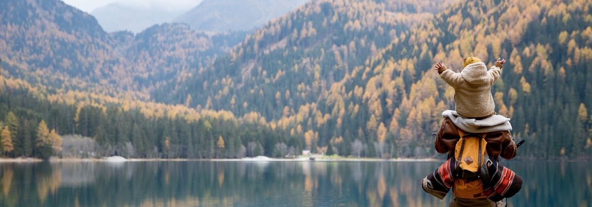 Ebeveyn omuzlarında göl kenarında sonbahar manzarasına bakan çocuk. Dağlar ve sarı ağaçlar huzurlu bir doğa kaçamağı sunuyor.