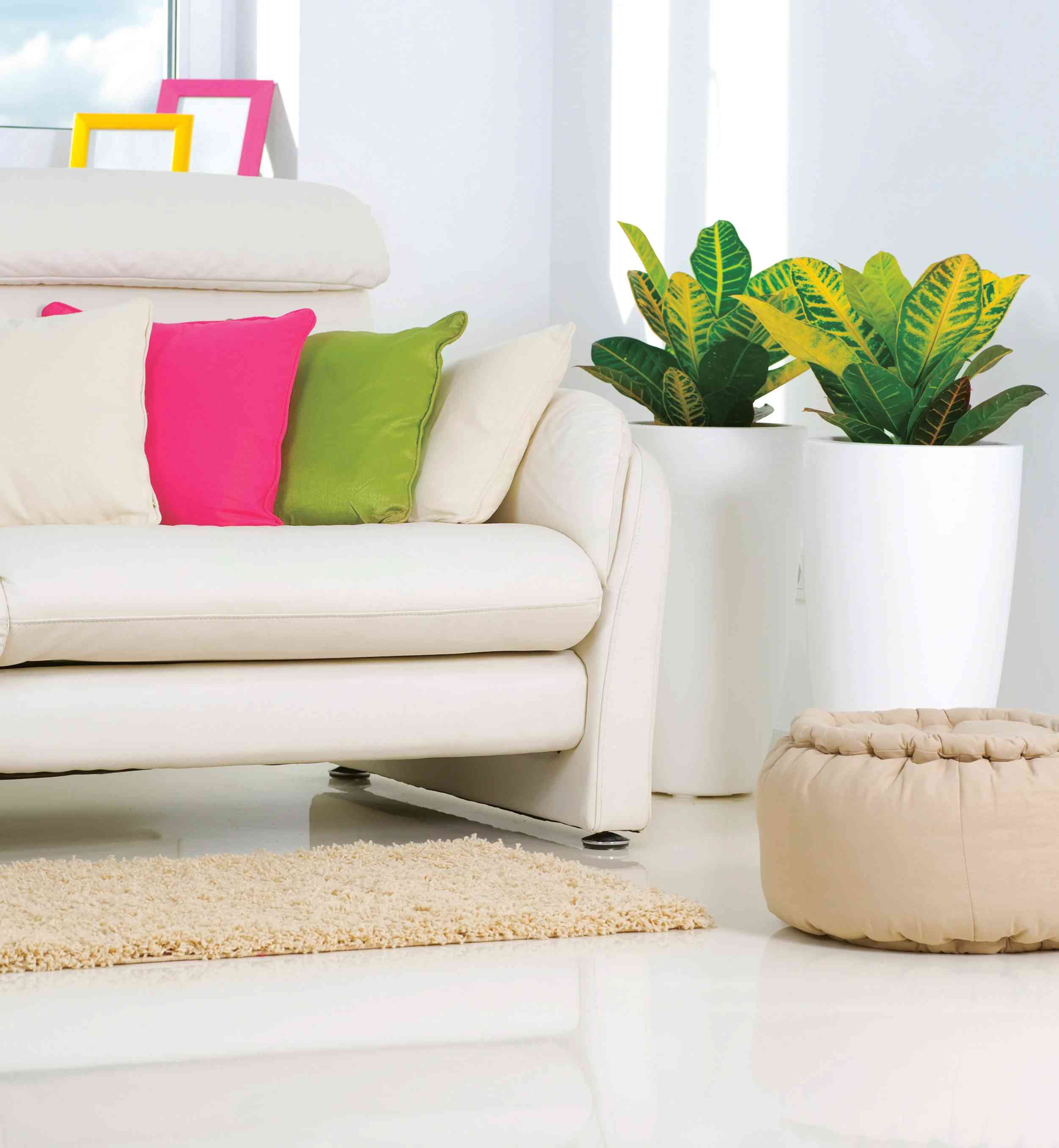 Modern, beyaz bir oturma odasında bej koltuk, renkli yastıklar, dekoratif bitkiler ve parlak zemin. Ferah ve şık iç mekan.