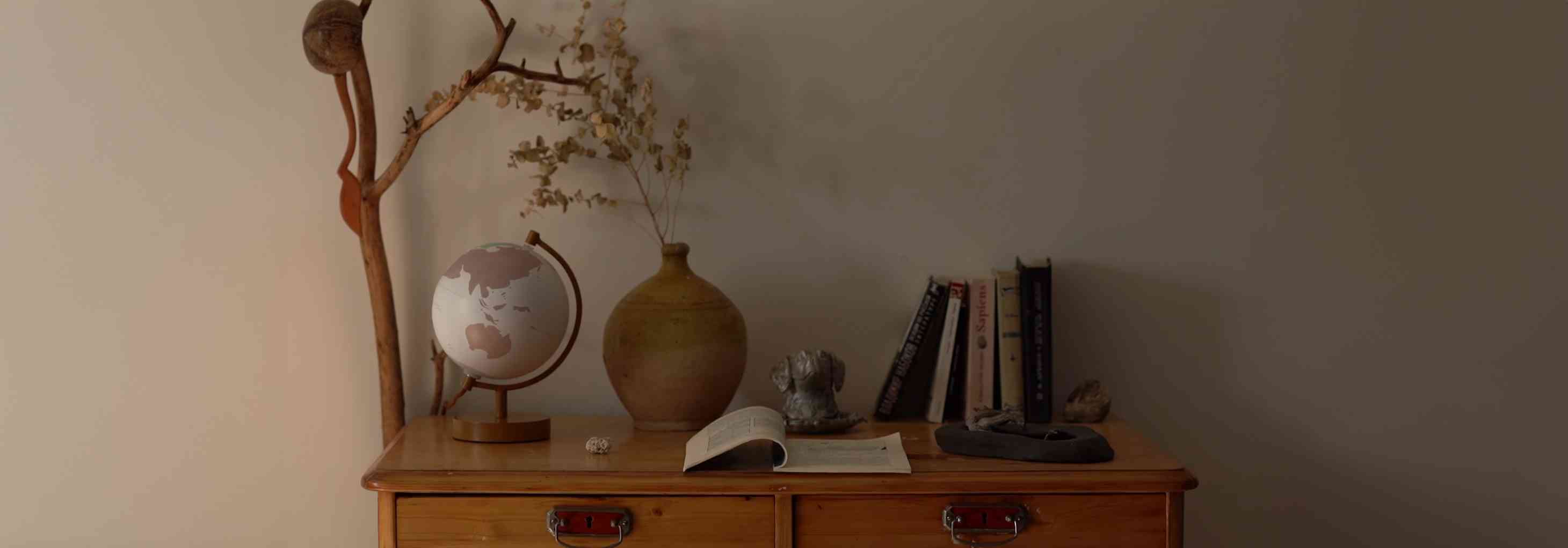 Ahşap komodin üzerinde dünya küresi, kitaplar ve kuru çiçekli vazo ile düzenli ev köşesi.